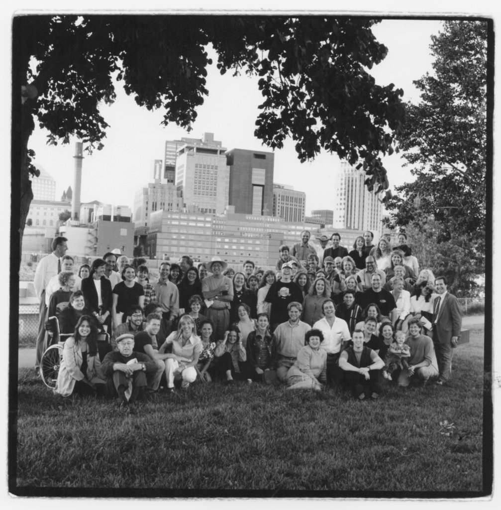 A black-and-white group photo of approximately 60 people gathered on a grassy area under tree branches. The group includes diverse individuals, some seated, some standing, and one person in a wheelchair. Everyone is smiling or posing together. In the background is a city skyline with tall buildings and industrial structures, suggesting an urban setting. The atmosphere appears friendly and communal.